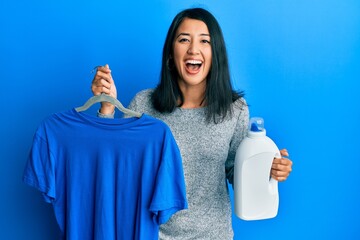 Beautiful asian young woman holding blue t shirt and detergent bottle smiling and laughing hard out loud because funny crazy joke.