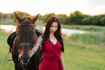 Obraz premium A young pretty girl in a red dress poses on a ranch with a thoroughbred stallion at sunset. Love and care for animals
