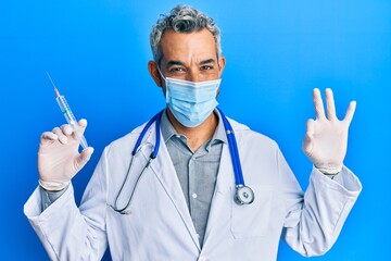 Middle age grey-haired man wearing doctor uniform and medical mask holding syringe doing ok sign with fingers, smiling friendly gesturing excellent symbol
