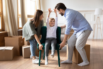 Happy young Caucasian parents carry in chair excited small son settle in own house. Smiling family with little boy child move furniture relocate to new house together. Realty, real estate concept.