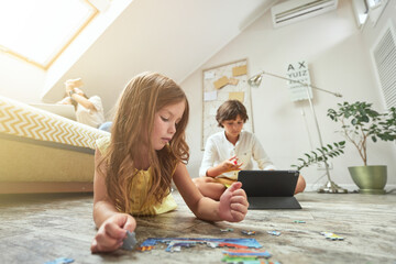 Family at home. Little girl lying on the floor in the living room and playing with puzzles while her brother using digital tablet and doing homework