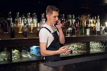 the bartender is talking on the phone at the bar against the background of bottles with a bar spoon in his hands.