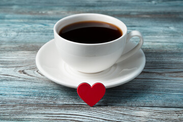 A small red heart is leaning against a coffee mug on a saucer on a wooden background. Concept of holiday backgrounds, February 14, love.