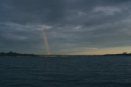 Far away rainbow under a cloudy sky over a large river in the amazonian forest in Brazil. Tropical vegetation on the horizon and sunlight shinning on the right as the sun sets. Space for text