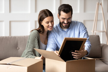 Smiling couple renters sit on sofa in new home unpack boxes with personal belongings moving together. Happy man and woman tenants unbox parcel relocate to own house. Relocation, rent concept.