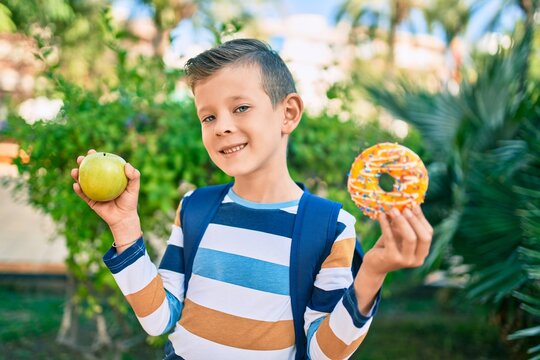 Adorable caucasian student boy smiling happy holding donut and bottle of water at the park.