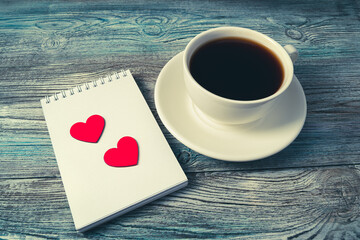 Two red hearts on a white Notepad next to a coffee Cup on a wooden background. Side view. Concept February 14.