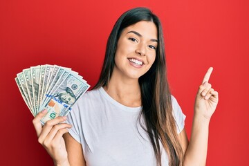 Young brunette woman holding dollars smiling happy pointing with hand and finger to the side