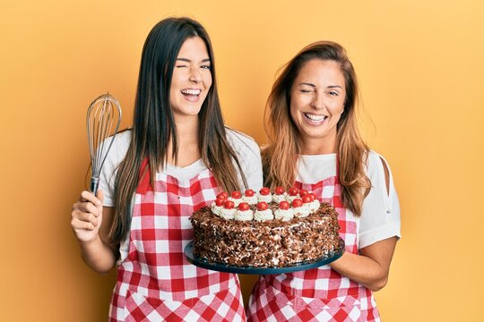 Hispanic family of mother and daughter wearing baker apron holding homemade cake winking looking at the camera with sexy expression, cheerful and happy face.
