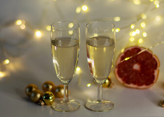 two glasses of champagne, pink grapefruit, golden glass balls and various Christmas decorations, illuminated background of a chain of white lights, Christmas waiting time