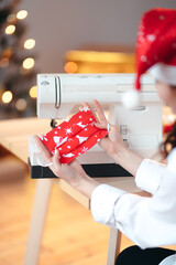 A girl in a red Christmas hat holds a green homemade medical mask with a Christmas print in her hands against the background of a festive tree and a sewing machine