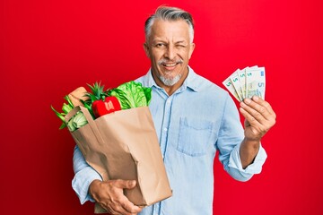 Middle age grey-haired man holding groceries and euro banknotes winking looking at the camera with...