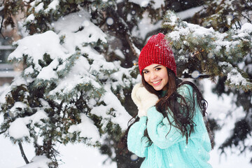 Fototapeta na wymiar an attractive young girl with dark flowing hair in a blue sweater, red hat and mittens smiles against the background of snow and Christmas tree branches in the frost.