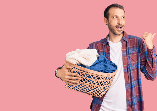 Young handsome man holding laundry basket pointing thumb up to the side smiling happy with open mouth
