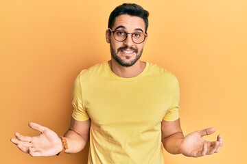 Young hispanic man wearing casual clothes and glasses clueless and confused expression with arms...