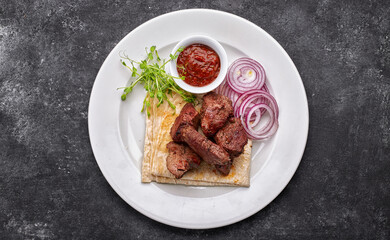 shashlik, with sauce, onions, and lavash, on a white plate, on a dark background