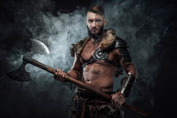 Fototapeta premium Barbaric scandinavian seafarer armed with two handed axe and dressed in fur with light armour holding huge axe in fog.