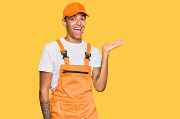 Young handsome african american man wearing handyman uniform smiling cheerful presenting and pointing with palm of hand looking at the camera.