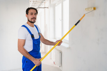 A bearded handyman in a blue overalls is painting a wall in the hallway. He uses a roller