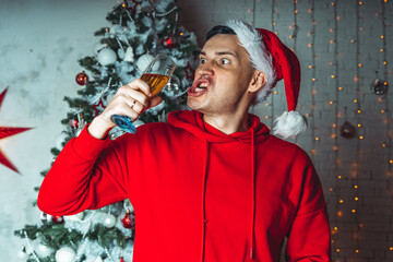 Young man in santa hat with glass of champagne on background of Christmas tree. Adult male posing at coniferous tree with decorative adornments. Concept of Christmas celebration at home.