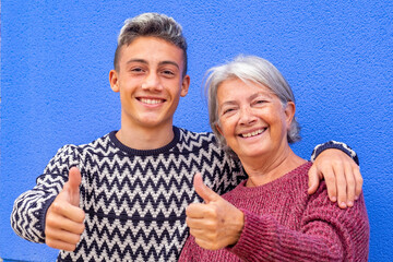 Happy family. Portrait of a white-haired grandmother and teenage grandson smiling and embracing looking at camera with thumbs up. Blue wall background