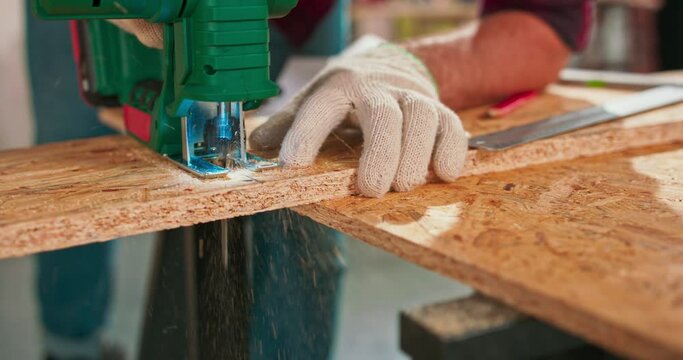 Young carpenter cuts an osb board with a jigsaw. Craftsman's hands in cloth protective gloves. The jigsaw blade cuts precisely.