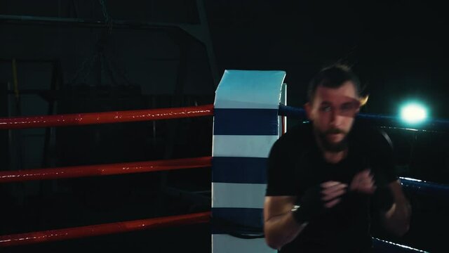 Boxer in dark sportswear with boxing wraps on fists looking at camera while practicing punches on boxing ring in the dark. Strong man working out in gym. Concept of sport