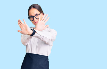 Beautiful brunette young woman wearing professional waitress apron rejection expression crossing arms and palms doing negative sign, angry face