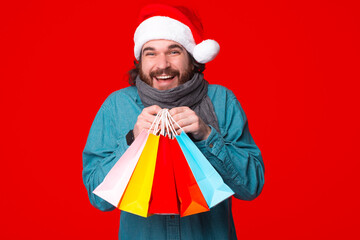 Bearded man holding shopping bags wearing santa hat standong over red background.