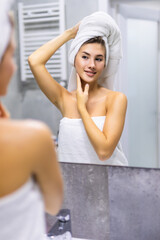 Young woman in bathrobe and towel on head looking at mirror in bathroom