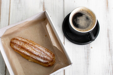 Eclair and coffee cup on a white wooden background.