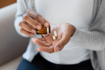 Crop close up of African American woman take pills or tablets from bottle. Biracial female have...