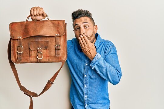 Handsome man with beard holding business bag covering mouth with hand, shocked and afraid for mistake. surprised expression