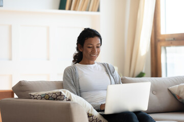 Fototapeta premium Smiling millennial African American woman rest sit on couch at home work online on laptop. Happy young biracial female relax on sofa in living room look at computer screen, browse wireless internet.