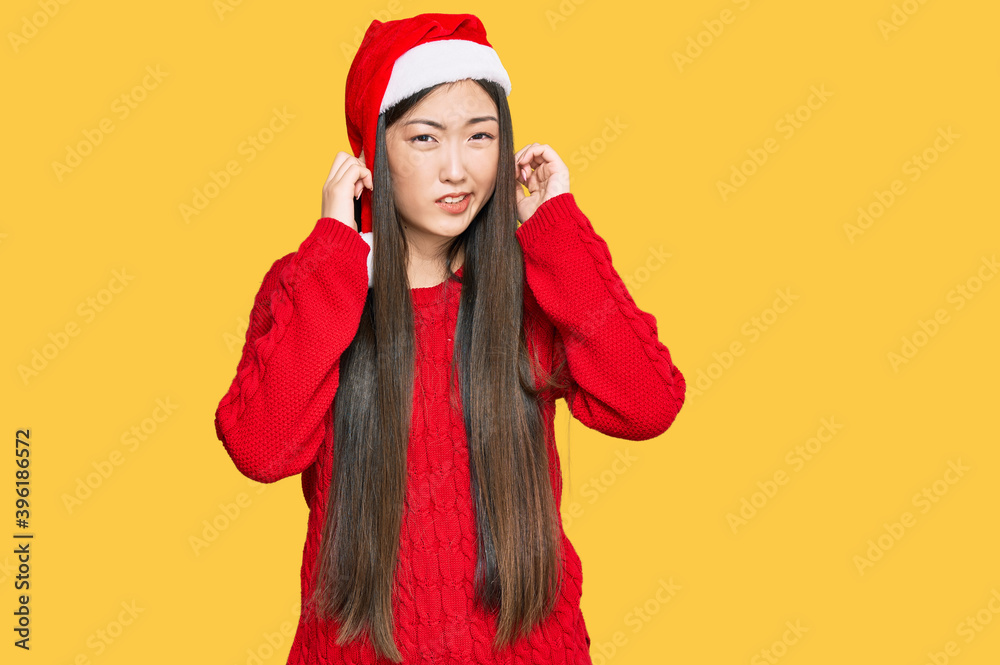 Sticker young chinese woman wearing christmas hat covering ears with fingers with annoyed expression for the