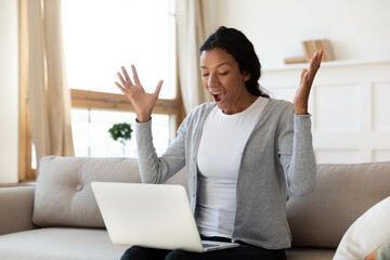 Excited young biracial woman sit on sofa at home look at laptop screen shocked by online discount...