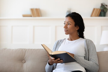 Young African American woman sit rest on sofa at home read book look in distance dreaming or thinking. Pensive dreamy biracial female enjoy novel lost in thoughts, relax on weekend in living room.