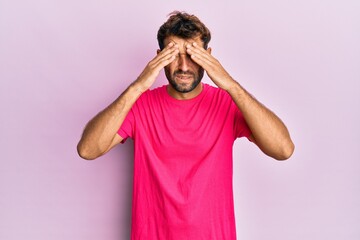 Handsome man with beard wearing casual pink tshirt over pink background suffering from headache desperate and stressed because pain and migraine. hands on head.