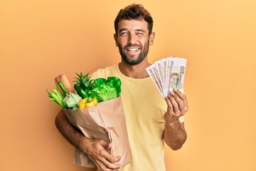 Handsome man with beard holding groceries and 100 ruble banknotes winking looking at the camera...