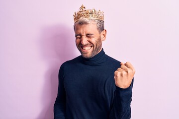 Young handsome blond man with piercing wearing golden king crown over pink background celebrating surprised and amazed for success with arms raised and eyes closed