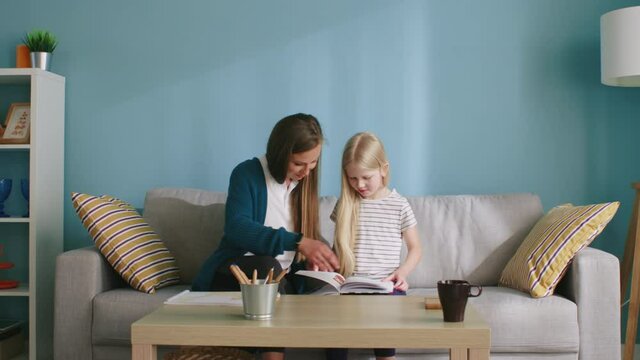 Young mom and her small girl are sitting on sofa and enjoying reading their favorite book, looking through the colorful pictures, enjoy their relationships, staying home, Slow motion.