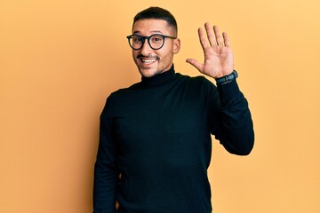 Handsome man with tattoos wearing turtleneck sweater and glasses waiving saying hello happy and...