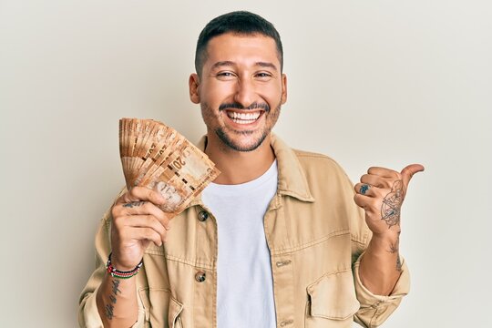 Handsome man with tattoos holding south african 20 rand banknotes pointing thumb up to the side smiling happy with open mouth
