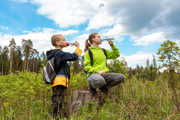 Teenager girl and little boy drink water from bottles while relaxing on forest trip. Tired hikers children with backpacks are rest