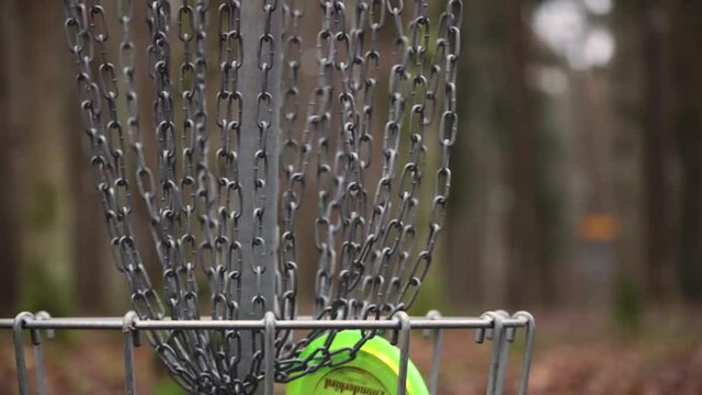 Disc Golf target basket yellow in autumn forest with moving chains and brown leaves fallen on the ground and trees in the background cloudy weather multiple scored discs close up moving chains slower
