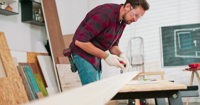 Portrait of hardworking professional carpenter holding a ruler and pencil while measuring a board in a carpentry workshop. A bearded DIY enthusiast measures wood. There are a locksmith table and tools
