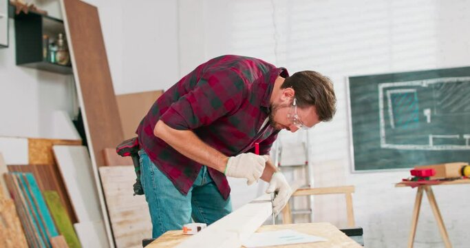 Portrait of hardworking professional carpenter holding a ruler and pencil while measuring a board in a carpentry workshop. A bearded DIY enthusiast measures wood. There are a locksmith table and tools