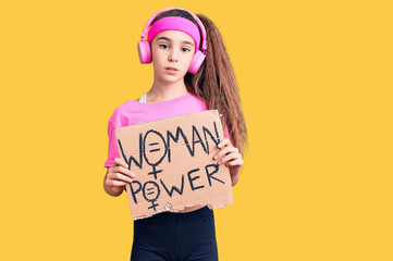 Cute hispanic child girl wearing gym clothes and using headphones holding woman power banner thinking attitude and sober expression looking self confident