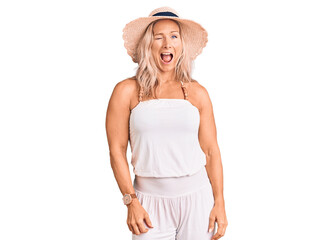 Middle age fit blonde woman wearing summer hat winking looking at the camera with sexy expression,...