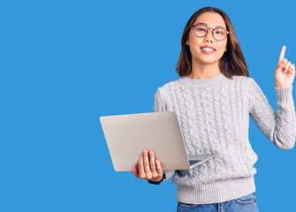 Young beautiful chinese girl wearing glasses holding laptop surprised with an idea or question pointing finger with happy face, number one